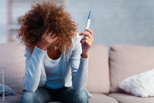 Depressed girl holding pregnancy test covering face with hand, sitting on couch, free space photo