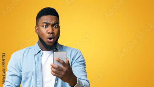 Unexpected message. Astonished african american man looking at smartphone screen in shock, standing over yellow background with copy space
