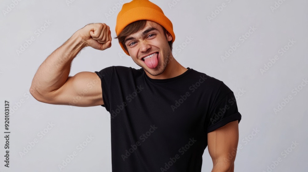 Naklejka premium Happy gen Z young man wearing black t-shirt and orange beanie flexing his bicep and sticking out tongue. Studio shot, white background