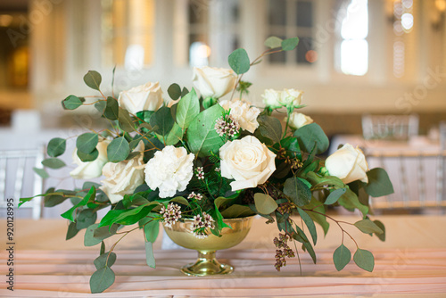 White roses and green leaf floral centerpiece in gold vase on table with bokeh background