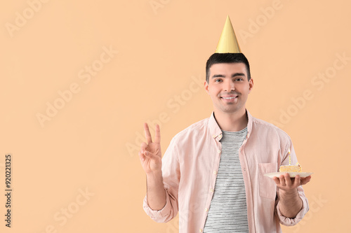 Young man with birthday cake showing victory gesture on beige background