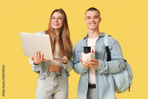 Young students with piggy bank and laptop on yellow background. Concept of sa...