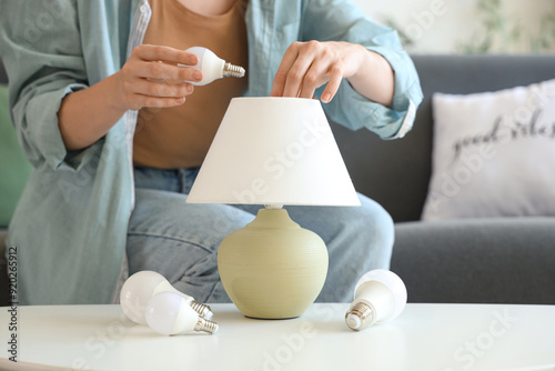 Fotografia Young woman screwing light bulb into lamp on table at home, closeup