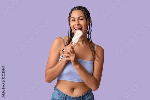 Beautiful young African-American woman eating sweet ice-cream on lilac background