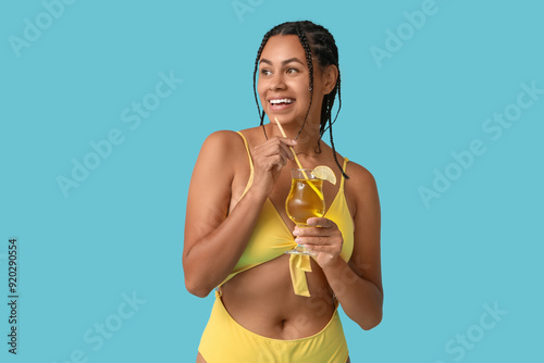 Beautiful young African-American woman in stylish yellow swimsuit with glass of cocktail on blue background