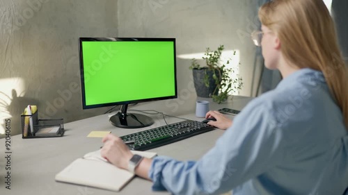 Young Woman Looks at Computer with Green Screen Mock Up Display and Writes Something in Notebook. Girl Working In Office On Computer With Green Screen