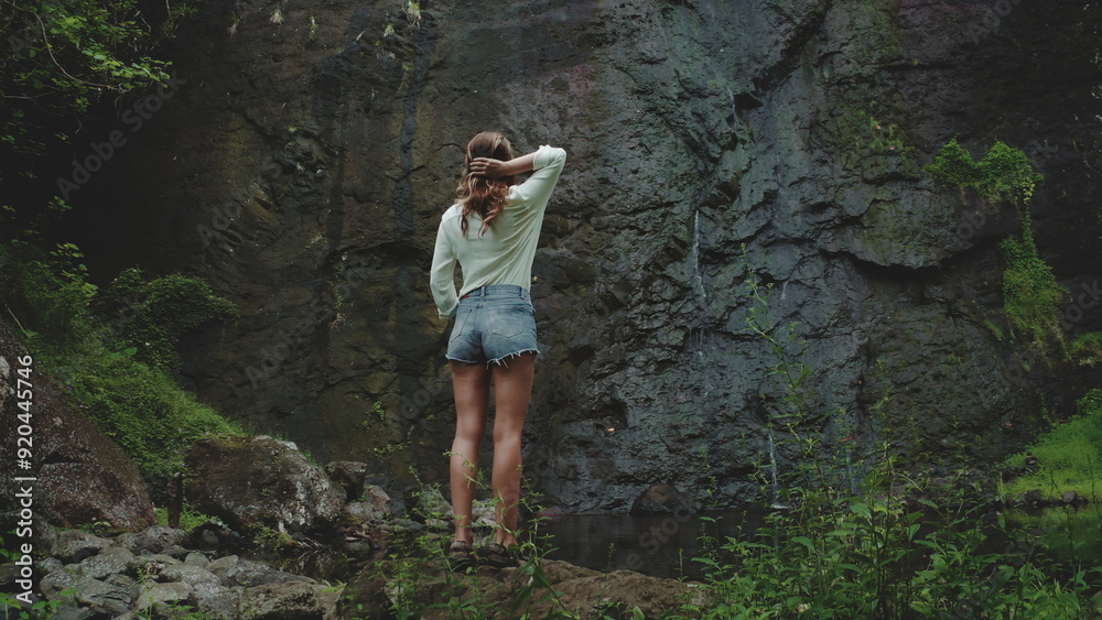 Naklejka premium Woman admires a hidden waterfall in a lush tropical forest, touching her hair in wonder. Perfect for summer adventures in french polynesia's moorea
