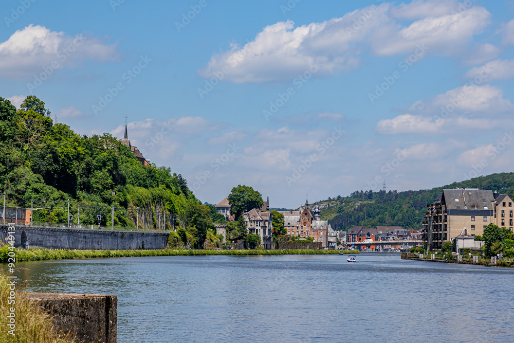 Obraz premium River Meuse with cityscape of Dinant resort town against blue sky in background, lush trees on hill, sunny summer day in Namur province, Wallonia, Belgium