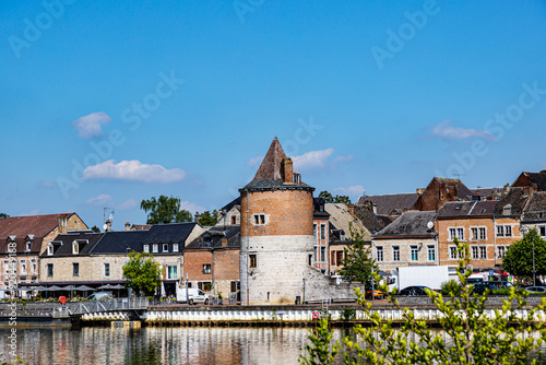 Medieval and ancient buildings in town of Givet against blue sky background, vehicular street, promenade and restaurant terraces along river Meuse, sunny summer day in Ardennes department, France