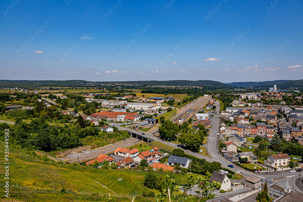 Obraz premium Aerial view of Givet city outskirts, bridge crossing railway tracks, vehicular streets, buildings, houses, trees in field and horizon in background, sunny summer day in Ardennes department, France