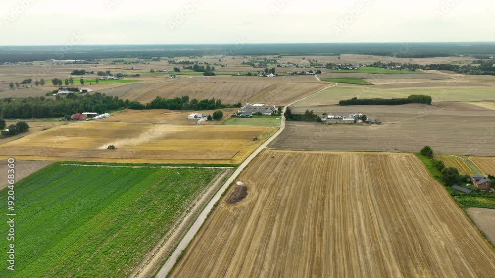 Aerial view of a patchwork of fields with a harvester in action. The fields are a mix of harvested and unharvested crops, with a road running through the landscape and farm buildings in the distance Aerial view of a patchwork of fields with a harvester in action. The fields are a mix of harvested and unharvested crops, with a road running through the landscape and farm buildings in the distance