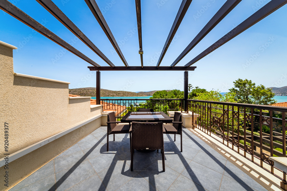Fototapeta premium Balcony or terrace with outdoor furniture under wooden beams meant for roof, overlooking garden and sea view. Space combines comfort and natural beauty. Perfect for relaxation and enjoying.