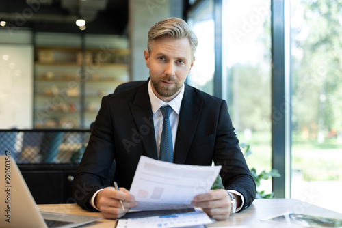 Mature businessman with beard working with documents, contracts and bills sitting at table using laptop at work in modern bright office with large windows