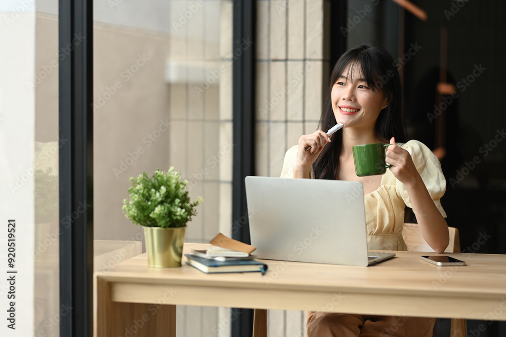 Charming young Asian woman staring out the window of office with a coffee cup in her hand Charming young Asian woman staring out the window of office with a coffee cup in her hand