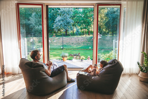 A little girl and her grandfather drinking tea, seated on bean bags, enjoying view of a lush garden through a large glass door