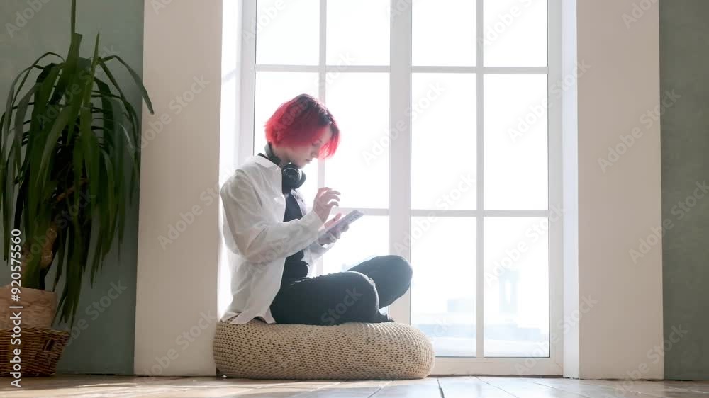 Sweet young Caucasian girl sits on an armchair near large bright window, puts on headphones during day and turns on music on her smartphone. girl listens to lectures online Sweet young Caucasian girl sits on an armchair near large bright window, puts on headphones during day and turns on music on her smartphone. girl listens to lectures online