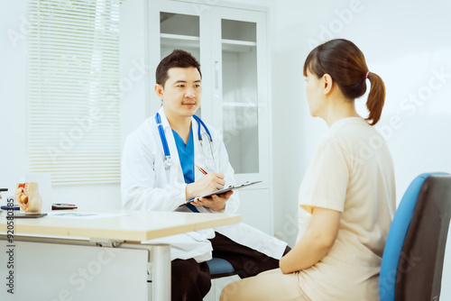 A male Asian pediatrician in a white lab coat sits at his desk, warmly greeting a middle-aged pregnant woman for a prenatal consultation, offering care and support in a clinical setting.