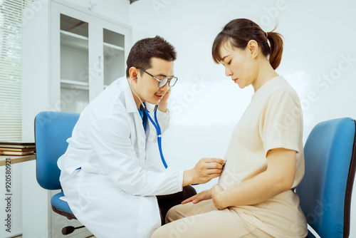 A male Asian pediatrician in a white lab coat sits at his desk, warmly greeting a middle-aged pregnant woman for a prenatal consultation, offering care and support in a clinical setting.