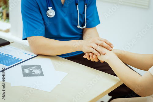 A male Asian pediatrician in white lab coat gently holds hand of middle-aged pregnant woman, encouragement and support during prenatal care consultation focused on maternal and child health. photo