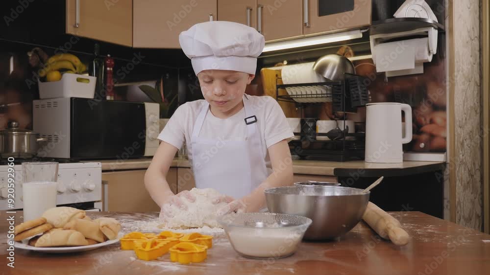 A little boy in a white apron and a chef's hat kneads the dough in the kitchen, there is a lot of flour and various ingredients on the table. A little boy in a white apron and a chef's hat kneads the dough in the kitchen, there is a lot of flour and various ingredients on the table.