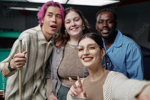 Group of friends enjoying time together at pool hall, taking selfie with happy expressions. Visible pool tables and cues in background creating relaxed atmosphere