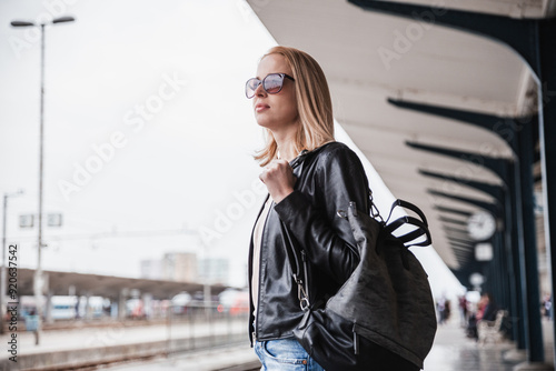 Young blond woman in jeans, shirt and leather jacket wearing bag and sunglass waiting for train to arrive at train station platform. Commuting, travel and transportation concept.