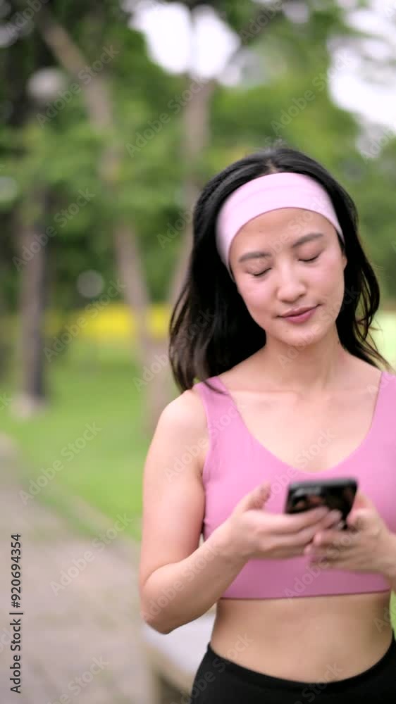 Vertical video of Asian woman in a pink sports top and headband is focused on smartphone, showing a look of concentration. She is outdoors, surrounded by greenery. Vertical video of Asian woman in a pink sports top and headband is focused on smartphone, showing a look of concentration. She is outdoors, surrounded by greenery.