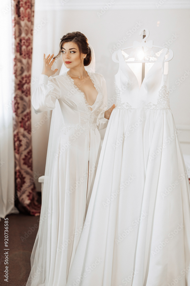 A bride is standing in front of two white wedding dresses. She is looking at the dresses and possibly trying to decide which one to wear A bride is standing in front of two white wedding dresses. She is looking at the dresses and possibly trying to decide which one to wear