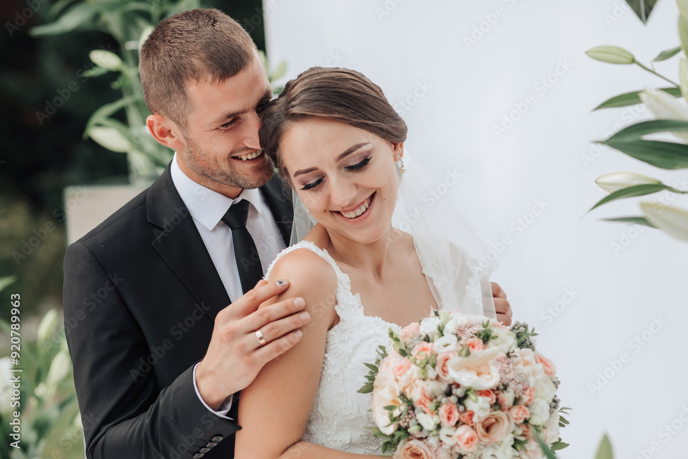 A bride and groom are hugging and smiling in front of a white background. The bride is holding a bouquet of flowers A bride and groom are hugging and smiling in front of a white background. The bride is holding a bouquet of flowers