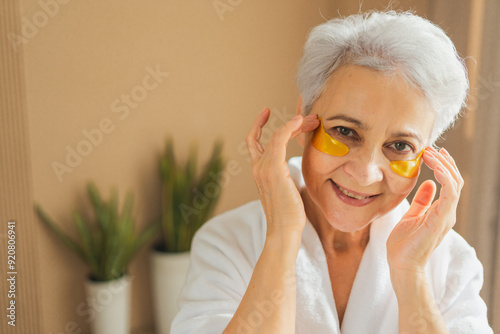 Eye patch beauty aging treatment. Mature adult old woman with under eye collagen pads. Senior woman with patches under eyes gets facial treatment. Senior lady in robe relaxing in bathroom at home
