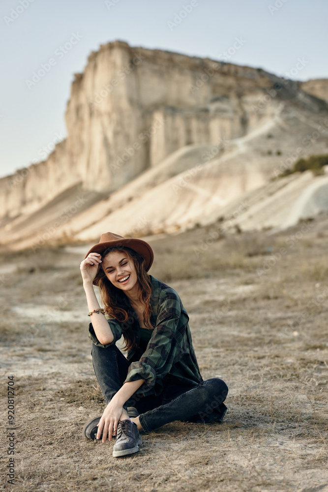 Serene woman in hat sitting on ground in front of majestic mountain with hands on knees Serene woman in hat sitting on ground in front of majestic mountain with hands on knees