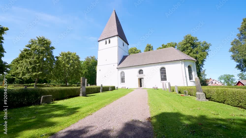 A serene white church with a tall steeple stands on the idyllic grounds of Bjalbo, Sweden, against a clear blue sky.