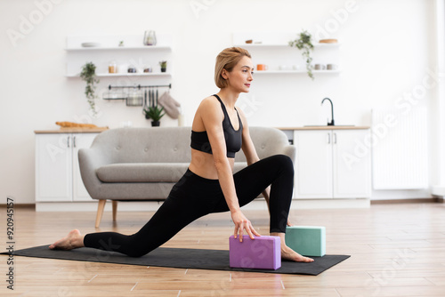 Woman practicing yoga indoors using blocks in modern living room setting, demonstrating flexibility and focus. Wearing black sportswear, she performs a low lunge pose on yoga mat.