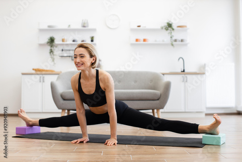 Woman practicing yoga splits on mat in bright home interior. Smiling and focused, performing flexible yoga pose using blocks for support on floor.