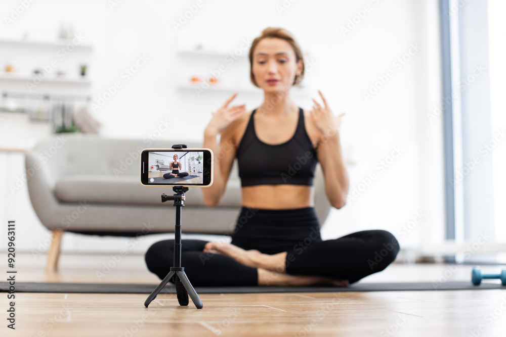 Yoga instructor sitting cross-legged on mat recording virtual fitness class using smartphone. Instructor demonstrating poses in modern living room. Concept of online fitness and digital wellness.