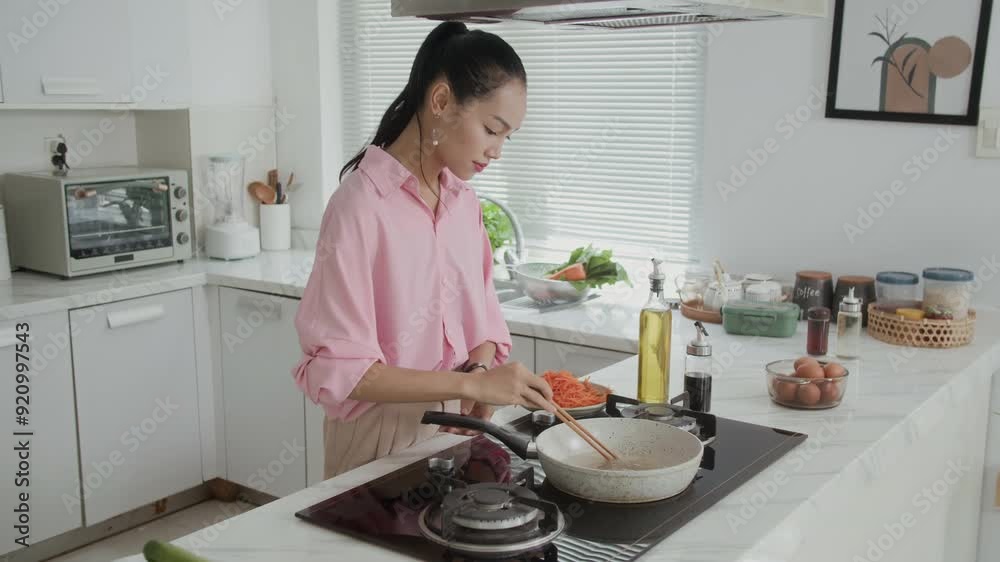 Medium long shot of young Asian girl frying sliced carrot on pan while cooking dinner at home kitchen Medium long shot of young Asian girl frying sliced carrot on pan while cooking dinner at home kitchen