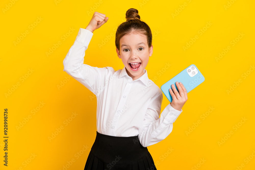 Photo of cute schoolgirl hold phone raise fist wear uniform isolated on yellow color background Photo of cute schoolgirl hold phone raise fist wear uniform isolated on yellow color background