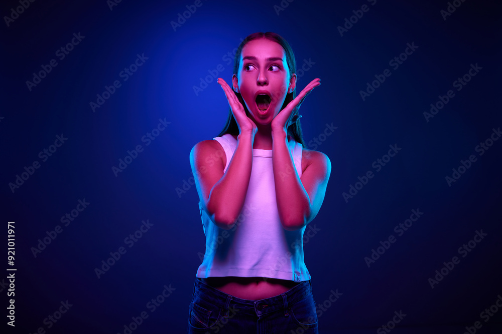 Beautiful young girl in casual white top expressing shock and positive excitement against gradient blue studio background in neon light. News, surprise, sales. Concept of human emotions, youth Beautiful young girl in casual white top expressing shock and positive excitement against gradient blue studio background in neon light. News, surprise, sales. Concept of human emotions, youth