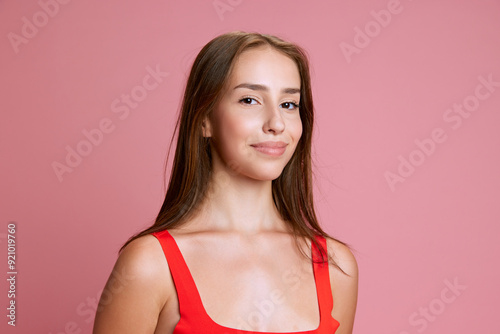 Portrait of beautiful young girl in red top posing with little tender smile against pink studio background. Concept of human emotions, beauty, fashion, youth, lifestyle