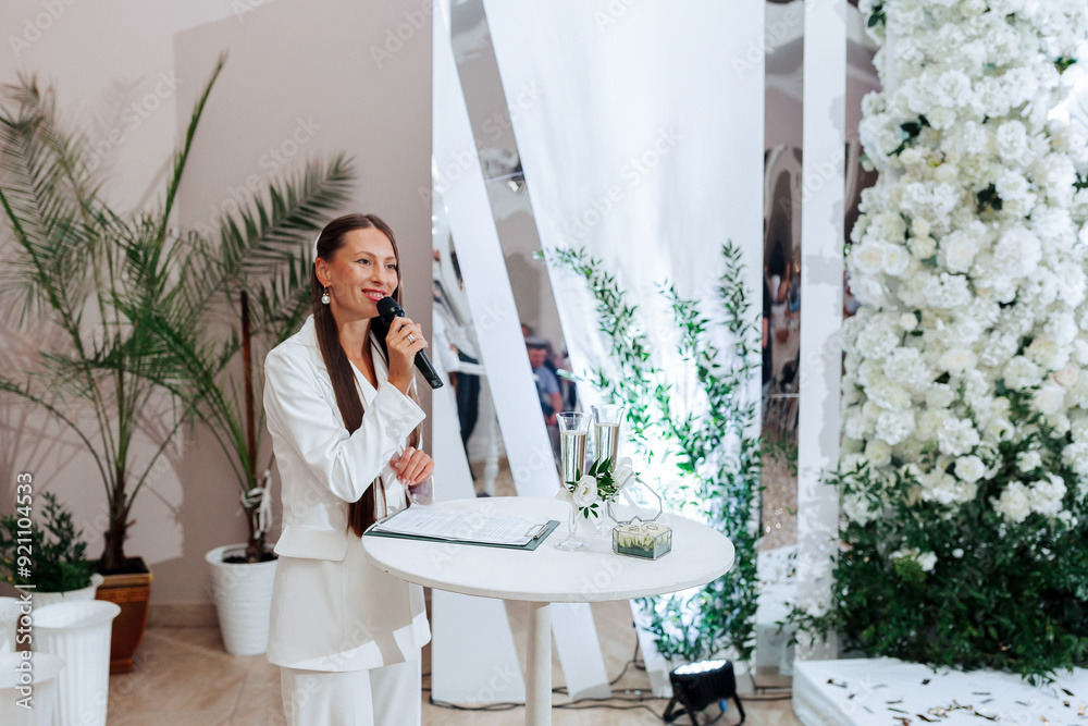 A woman is standing in front of a table with a microphone and a white background. She is speaking into the microphone A woman is standing in front of a table with a microphone and a white background. She is speaking into the microphone
