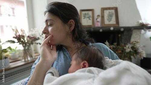 Obraz na plátně Thoughtful mother holding newborn baby, gazing out of window, seated in a comfor