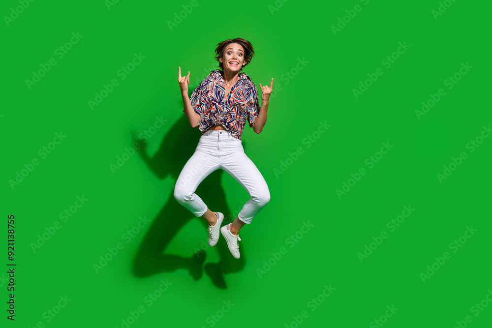 Full size photo of overjoyed girl wear print shirt jumping at rock concert showing horns symbol isolated on green color background Full size photo of overjoyed girl wear print shirt jumping at rock concert showing horns symbol isolated on green color background