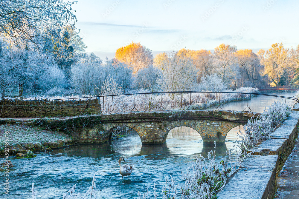 The old stone bridge over the River Coln and hoar frost on Rack Isle at sunset in the Cotswold village of Bibury, Gloucestershire, England UK
