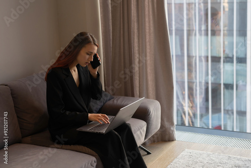 Young beautiful woman in a business suit sitting on a sofa by panoramic window in skyscraper apartment with a laptop and talking on a mobile phone. Remote working, freelance. Modern technology