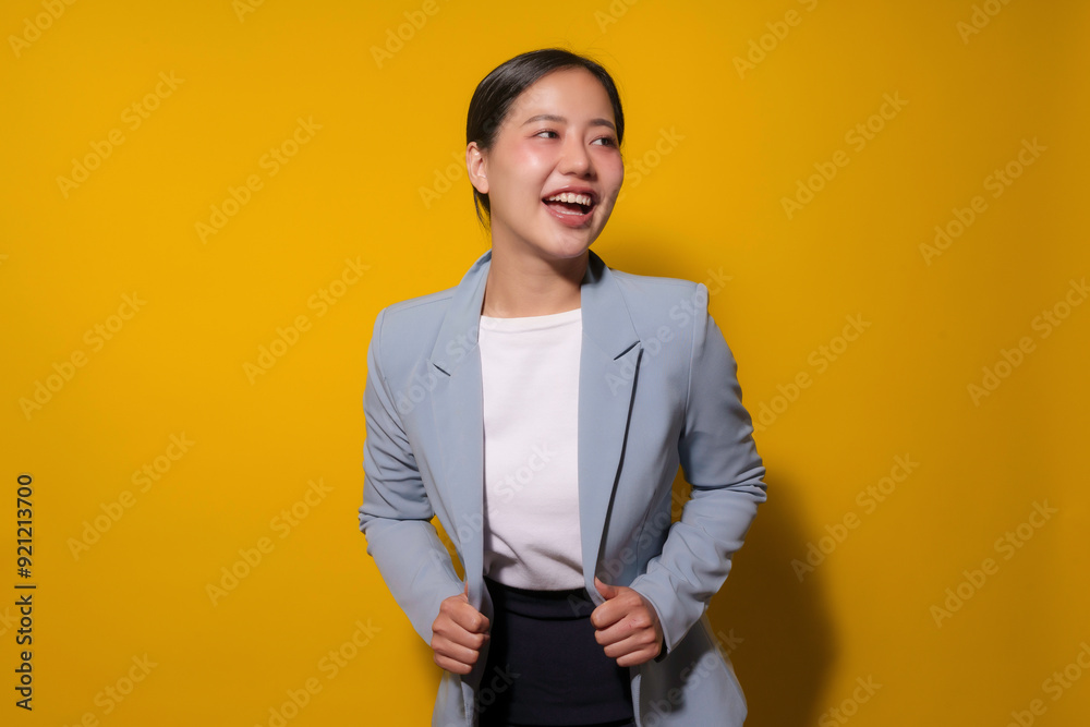 Young professional woman is standing against a yellow background, wearing a blue blazer and smiling while looking away Young professional woman is standing against a yellow background, wearing a blue blazer and smiling while looking away