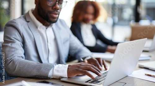 A focused professional is using a laptop in a bright office environment, with another colleague visible in the background, dressed in formal business attire, working diligently.