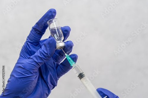 Close-up of hands wearing blue latex gloves holding a vial and using a syringe to extract its contents. The background is out of focus with a neutral grey tone