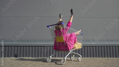 Beautiful woman in pink costume having fun in the grocery cart under the supermarket outdoors. Women racing shopping trolleys on the street background. Positive female going crazy