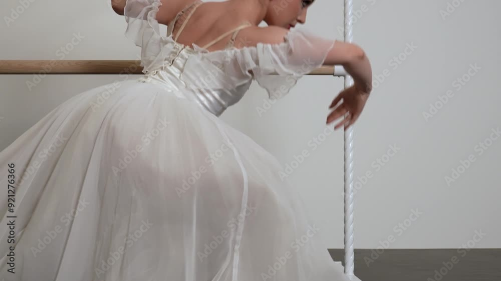 A beautiful ballerina in a white dress warming upi n a white sunny room with a black floor next to a ballet barre.