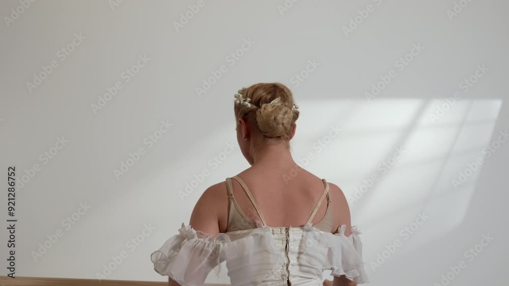 A beautiful ballerina in a white dress warming upi n a white sunny room with a black floor next to a ballet barre.